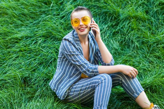 Portrait of beautiful young woman with short hair in casual blue striped suit, yellow shirt and glasses sitting on green grass on talking on her mobile phone and toothy smile. outdoor summertime shot.