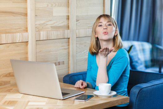 Portrait of lovely romantic young girl freelancer with blonde bob haircut hair in blue blouse are sitting alone in cafe and working on laptop and sending air kiss, looking at camera.
