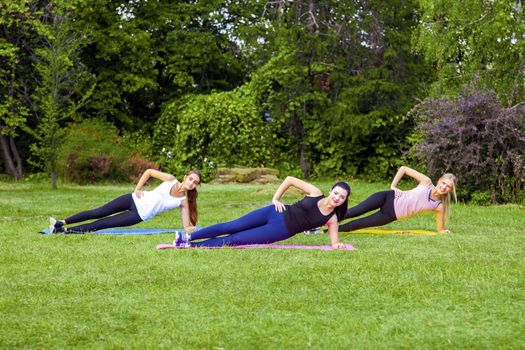 Group of three beautiful healthy slimy woman doing exersices on the green grass in the park, side palnk, looking at camera with toothy smile. Outdoor morning, sport lifestyle wellness and weight loss.