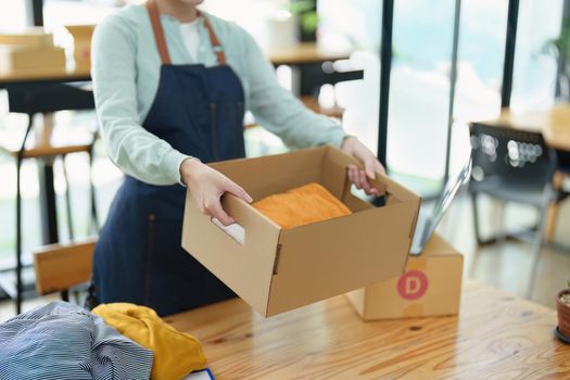 Portrait of a small startup Asian female entrepreneur SME owner picking up a yellow shirt before packing it in an inner box with a customer. Online Business Ideas and Freelance.