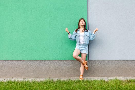 Full length portrait of beautiful woman in casual jeans denim style in summertime standing near green and light blue wall and doing yoga meditating with closed eyes and raised arms.
