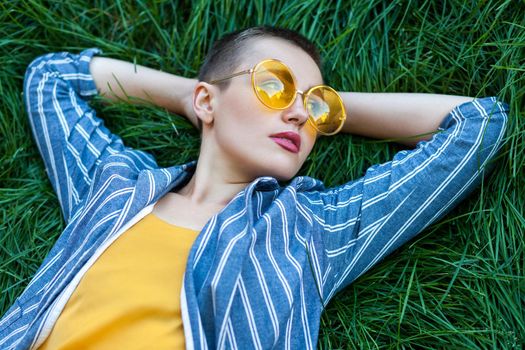 Portrait of thoughtful young woman with short hair in casual blue striped suit, yellow shirt, glasses lying down on green grass, holding hands behind head, looking, thinking. outdoor summertime shot.
