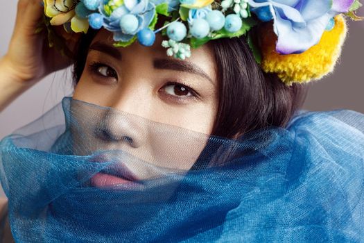 portrait of asian woman with floral hat and blue veil looking at camera on light grey background . indoor studio shot.