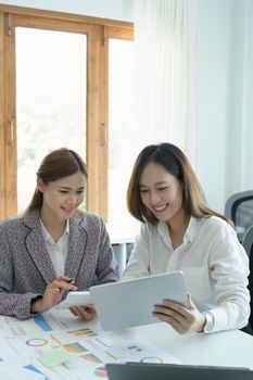 A portrait of two young Asian businesswoman using their tablet computers with calculators and documents to plan marketing strategies and investments to profit from their clients.