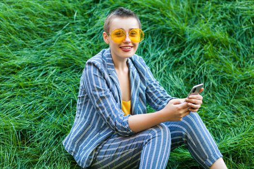 Portrait of pretty young short hair woman in casual blue striped suit, yellow glasses sitting on grass holding her smart phone mobile and looking at camera with toothy smile. outdoor summertime shot.