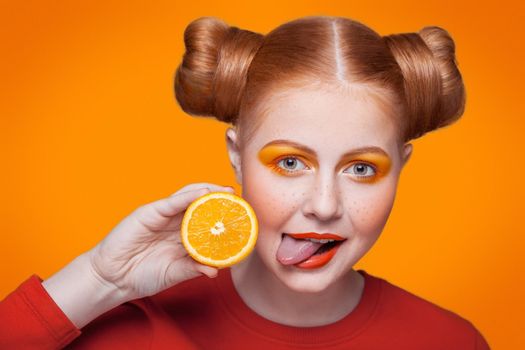 Young beautiful funny fashion model with orange slice on orange background. with orange makeup and hairstyle and freckles. studio shot, looking at camera and shows her tongue.