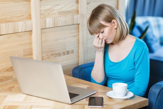 Side view portrait of sad young girl freelancer with blonde bob haircut hair in blue t-shirt are sitting in cafe and working on laptop,closed eyes and trying to stop crying hold fingers on the eyes