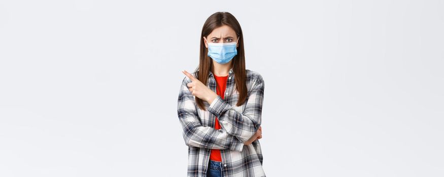 Coronavirus outbreak, leisure on quarantine, social distancing and emotions concept. Angry and disappointed young female in medical mask, pointing finger upper left corner, frowning mad.