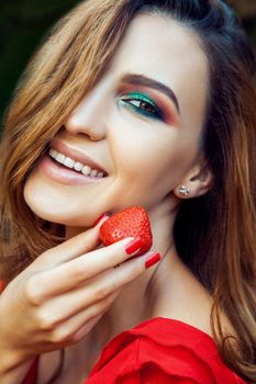 Young beautiful happy funny girl with red dress and makeup holding strawberry in summertime in the park. healthy lifestyle, diet beauty and happiness concept.