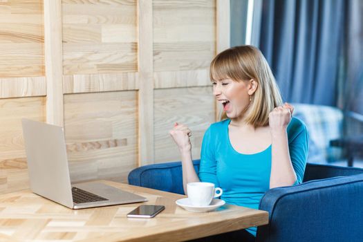 Side view portrait of beautiful amazed satisfied positive young girl freelancer with blonde hair in blue t-shirt are sitting in cafe and watching online reality show with toothy smile and triumphing