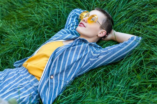 Portrait of dreamy beautiful young woman with short hair in casual blue striped suit, yellow shirt and glasses lying down on green grass, resting, dreaming and looking at sky. outdoor summertime shot.