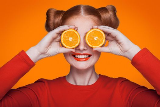 Young beautiful funny fashion model with orange slice on orange background. with orange makeup and hairstyle and freckles. holding orange between eyes with toothy smile.