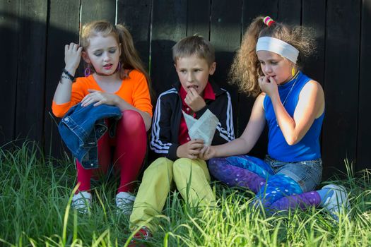 Cheerful kids, a boy and two cool girls sit on the grass near a wooden fence.