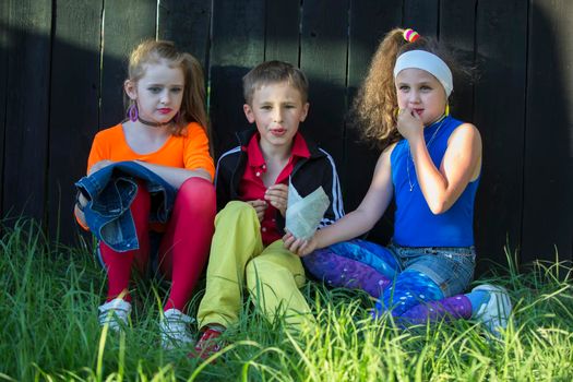 Beautiful model children in bright clothes sit on the grass near a wooden fence.