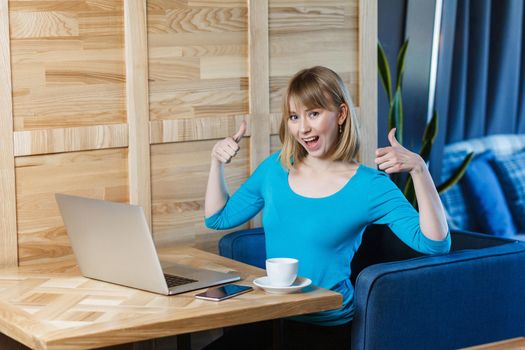 Portrait of attractive positive young girl freelancer with blonde bob haircut hair in blue blouse are sitting in cafe and working on laptop with toothy smile and showing thumb up, looking at camera.