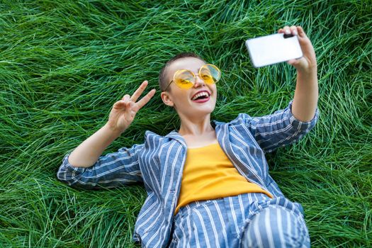 Happy young short hair woman in casual blue striped suit, yellow shirt, glasses lying down on green grass with mobile phone, doing selfie or online video with peace victory, excited. summertime shot.