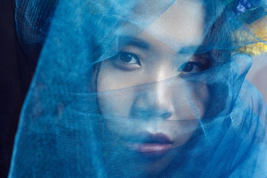 portrait of sensual gorgeous asian woman looking at camera through blue veil on face . indoor studio shot.