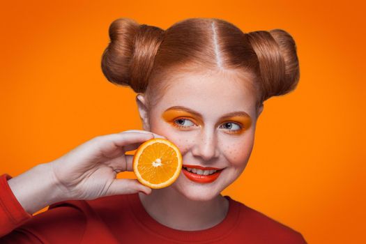 Young beautiful funny fashion model with orange slice on orange background. with orange makeup and hairstyle and freckles. studio shot, side looking with smile.