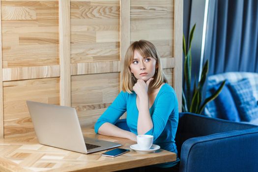 Beautiful thinkful young girl freelancer with blonde bob haircut hair in blue blouse are sitting in cafe and working on laptop, having new idea and planning own strategy, holding one hand on chin.