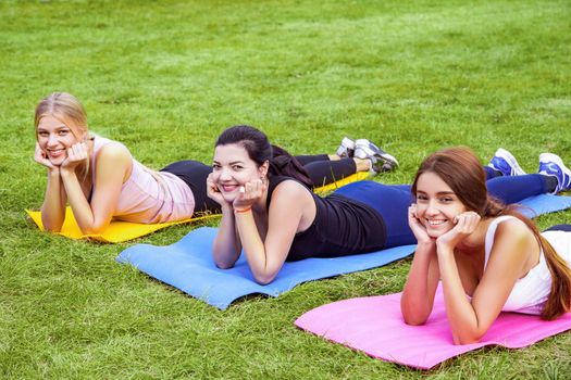 Group of beautiful satisfied healthy slimy young friends have rest after doing exersices on the green grass in park, lie on mat and head up with hands. Outdoor, sport lifestyle, yoga time, wellness