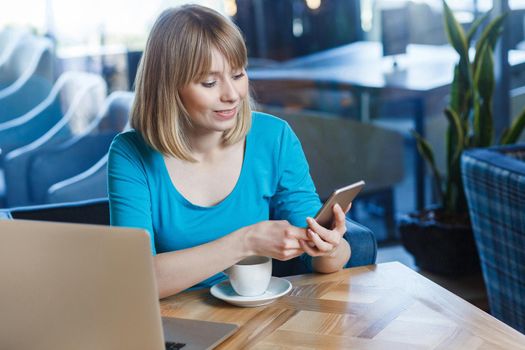 Top view portrait of young happy blonde woman in blue t-shirt, sitting in cafe and holding her mobile smart phone and texting. indoor studio shot.