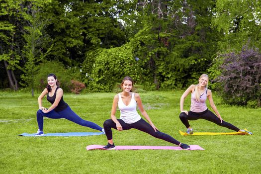 Group of beautiful healthy slimy woman doing exersices on mat in the green grass in the park, streching legs, looking at camera with toothy smile. Outdoor, happiness and spory lifestyle, morning