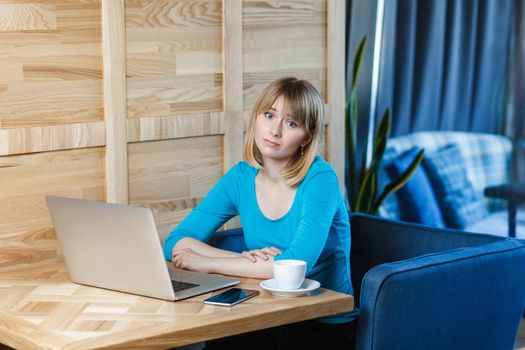 Dont know! Portrait of sadness tired young girl freelancer with blonde bob haircut hair in blue t-shirt are sitting alone in cafe and searching for a work on laptop are in bad mood without good result