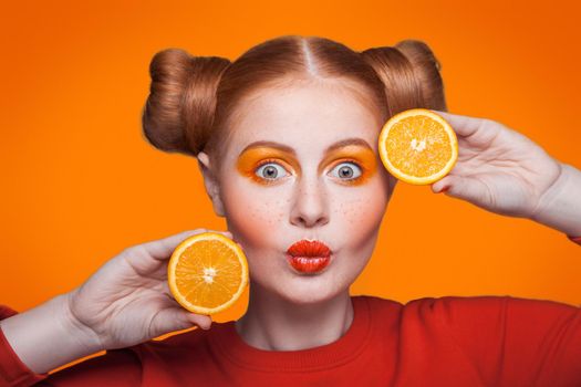 Young beautiful funny fashion model with orange slice on orange background. with orange makeup and hairstyle and freckles. studio shot, looking at camera with kiss.