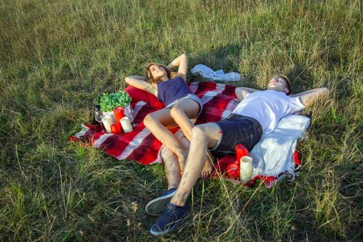 Young beautiful lovely couple lying down and have a rest on plaid in field on summer day and smiling.
