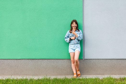 Full length portrait of happy excited beautiful woman in casual jeans denim style in summertime standing near green and light blue wall and pointing at green copyspace. two different choice concept.