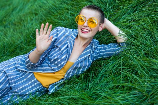 Portrait of beautiful young woman with short hair in casual blue striped suit, yellow shirt glasses lying down on green grass, looking at camea with toothy smile and greeting. outdoor summertime shot.