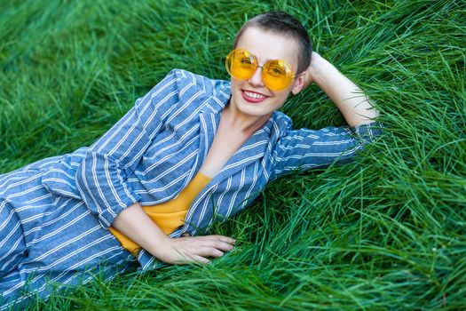 Portrait of beautiful young woman with short hair in casual blue striped suit, yellow shirt and glasses lying down on green grass, relax, looking at camera with toothy smile. outdoor summertime shot.