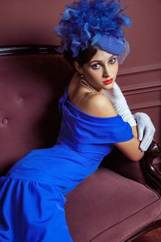 Old Fashioned retro great britain style photography. Beautiful young caucasian model in blue dress and fashion makeup and hat posing sitting on the artificial sofa and looking into the distance.