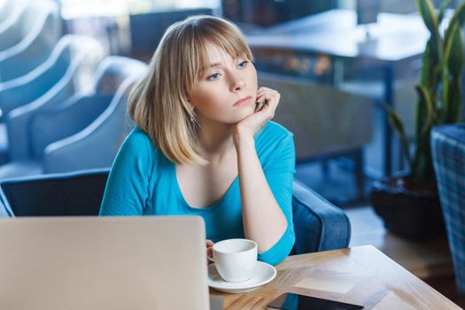 Beautiful thinkful young girl freelancer with blonde bob haircut hair in blue blouse are sitting in cafe and dreaming, having new idea and planning own strategy, holding one hand on chin.