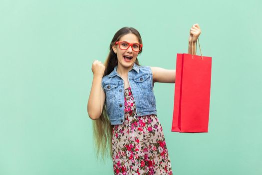 The beautiful long haired girl in casual clothing with shopping bags, looking at camera, celebrates the victory. Blonde model. Sales, shop, retail, consumer concept. Isolated studio shot on light blue background