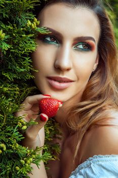 Closeup portrait of woman with perfect skin and makeup, holding near face red sweet strawberry. Outdoor spring or summer photo