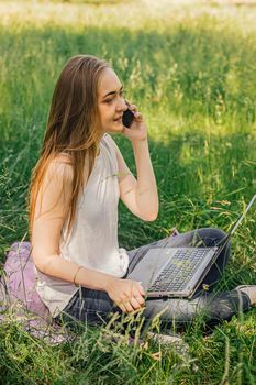 girl sits on the grass and works at a laptop. talking on the phone. freelance. selfeducation. the concept of remote learning and outdoor work