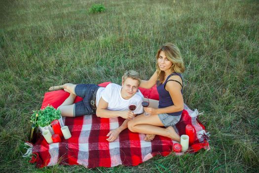 Beautiful young happy loving couple on picnic lying down on plaid in field on sunny summer day enjoying, holding and drinking wine and resting. looking at camera and smiling. top view.