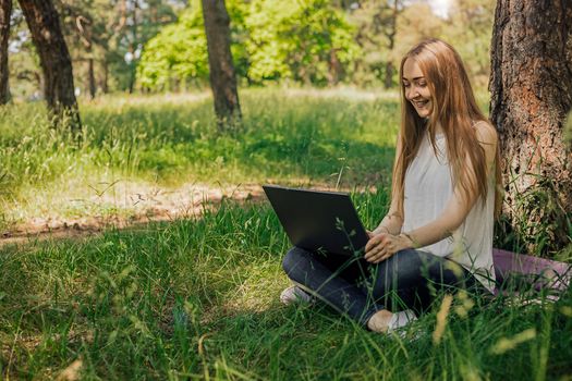 On the banner, a young girl works with a laptop in the fresh air in the park, sitting on the lawn. The concept of remote work. Work as a freelancer. The girl takes courses on a laptop and smiles