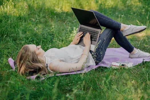 On the banner, a young girl works with a laptop in the fresh air in the park, sitting on the lawn. The concept of remote work. Work as a freelancer. The girl takes courses on a laptop and smiles