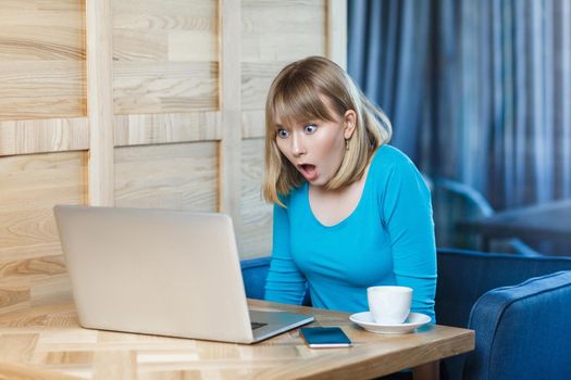 Don`t believe! Side view portrait of emotional shocked young businesswoman in blue t-shirt are sitting in cafe, reading news and remotely working with suprised big eyes looking to the laptop. Indoor