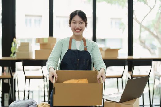 Portrait of a small startup Asian female entrepreneur SME owner picking up a yellow shirt before packing it in an inner box with a customer. Online Business Ideas and Freelance.