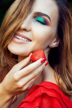 Young beautiful happy funny girl with red dress and makeup holding strawberry in summertime in the park. healthy lifestyle, diet beauty and happiness concept.