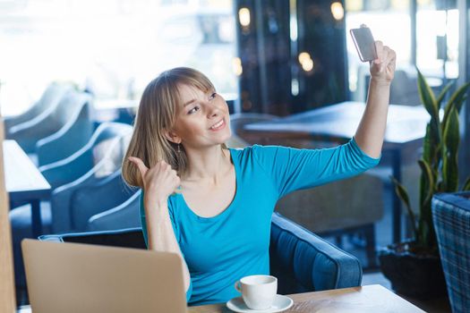 Portrait of happy satisfied funny young blonde woman in blue shirt holding mobile smart phone, making selfie or video call with toothy smile and thumbs up. indoor studio shot.