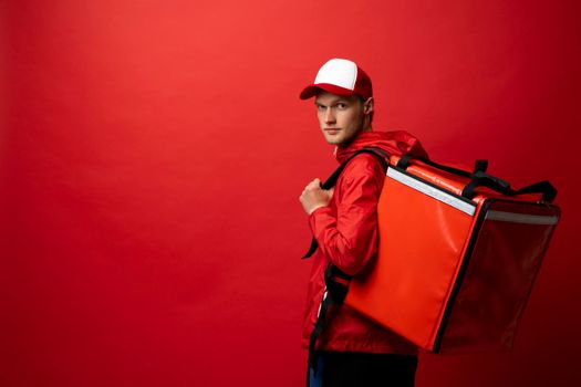 Delivery guy employee man in red cap T-shirt uniform work as dealer courier hold thermal food bag backpack isolated on red background