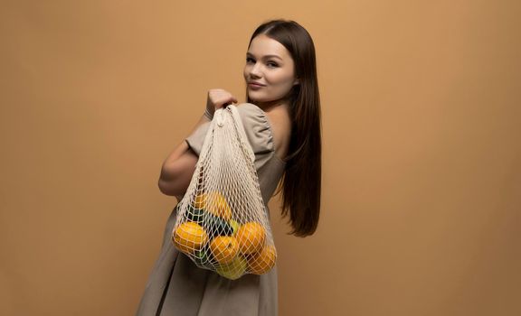 Woman in beige dress holding string shopping reusable mesh bag with vegetables, fruits on a shoulder. Zero waste concept. Space for text. Reusable eco friendly object. Copy space