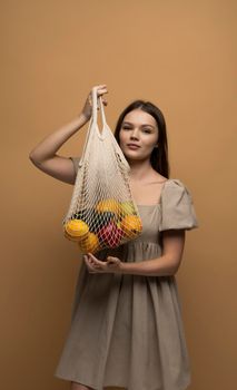 Eco friendly lifestyle and healthy diet concept, zero waste. Brunette woman in beige dress holding reusable mesh shopping bag with fresh fruits and vegetables