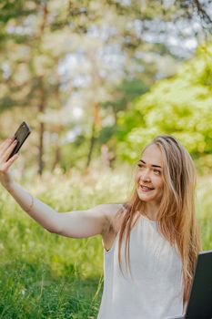 girl sits outdoors and works at a laptop. makes a selfie on the phone. freelance. selfeducation. the concept of remote learning and outdoor work