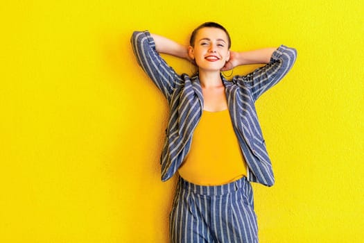 Portrait of happy young short hair beautiful woman in yellow shirt and striped suit standing, holding her head and looking at camera with toothy smile. indoor studio shot isolated on yellow background