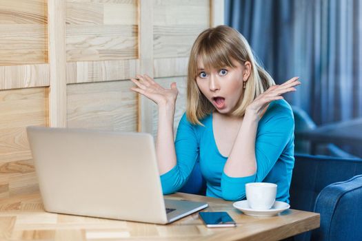 Wow! Portrait of emotional shocked young businesswoman in blue blouse are sitting in cafe, reading news and remotely working with suprised big eyes, open mouth and raised arms looking at camera.Indoor